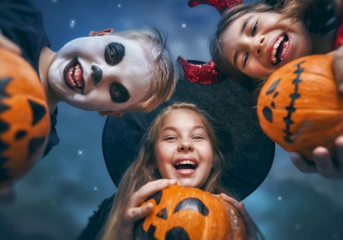 Three kids in Halloween costumes, with face paint and props, hold carved pumpkins and smile at the camera against a night sky.