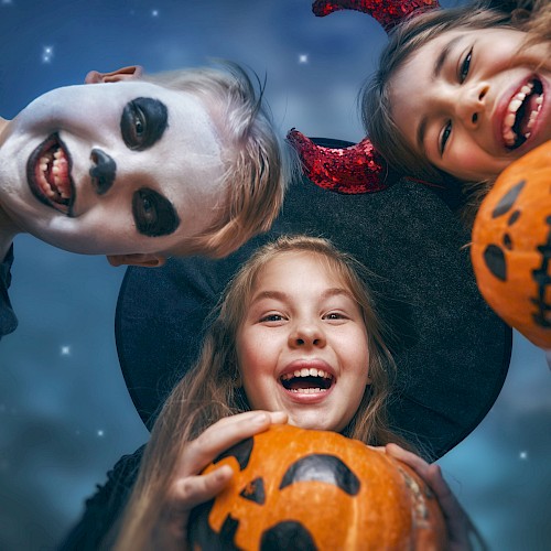 Three kids in Halloween costumes, with face paint and props, hold carved pumpkins and smile at the camera against a night sky.