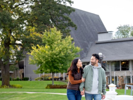 A couple is walking hand in hand through a park with trees and a modern building in the background.