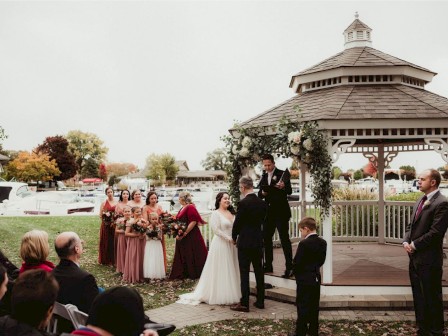 A wedding ceremony takes place outdoors by a gazebo, with a couple, officiant, wedding party, and guests in attendance.