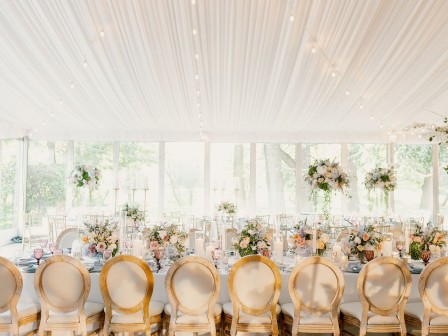 An elegant event setup with a long table, adorned with flowers and candles, under a draped ceiling with string lights, in a bright venue.