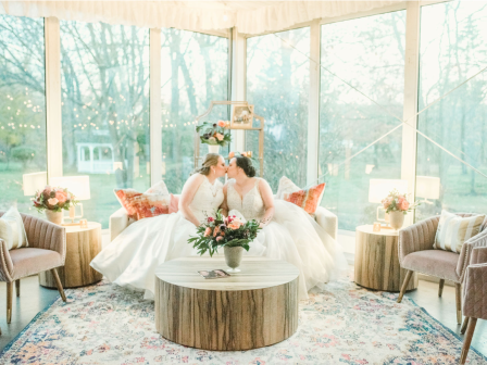 Two brides in wedding dresses are kissing on a sofa in a beautifully decorated room with flowers and elegant furniture.