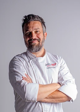 A confident chef with a grey beard and arms crossed, wearing a white chef&rsquo;s jacket against a plain backdrop.