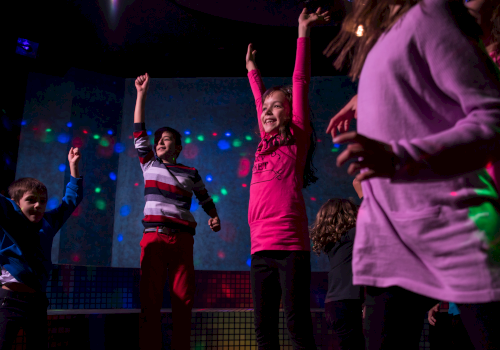 Children are dancing joyfully in a room with colorful lights, some with arms raised, wearing casual clothes.
