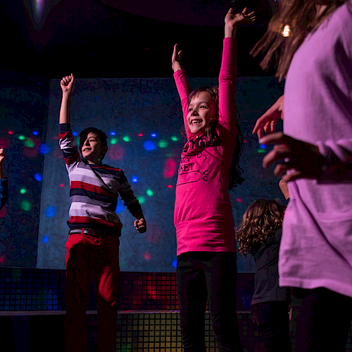Children are dancing joyfully in a room with colorful lights, some with arms raised, wearing casual clothes.